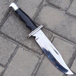 May include: A large, stainless steel knife with a black handle and a silver bolster. The blade is highly polished and reflects the surroundings. The knife is designed for outdoor use and has a sturdy construction.
