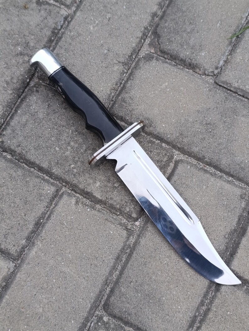 May include: A large, stainless steel knife with a black handle and a silver bolster. The blade is highly polished and reflects the surroundings. The knife is designed for outdoor use and has a sturdy construction.