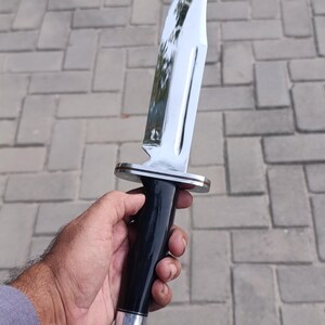 May include: A large, polished hunting knife with a shiny, curved blade. The handle is black with a silver bolster and pommel. The knife is held in a hand against a gray brick background.
