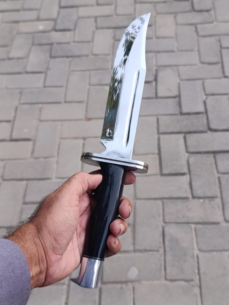 May include: A large, polished hunting knife with a shiny, curved blade. The handle is black with a silver bolster and pommel. The knife is held in a hand against a gray brick background.