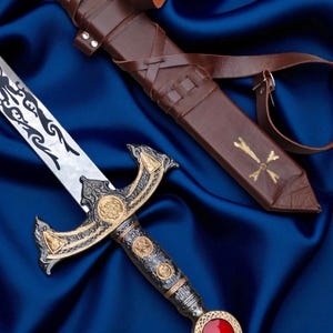 Op de afbeelding: Een sierlijk zwaard met een zilveren lemmet en decoratieve zwarte etsen. De pareerstang heeft gouden en zwarte accenten, met een rode edelsteen op de pommel. Een bruine leren schede met een kruisontwerp is ook aanwezig.