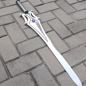 May include: A silver-colored sword with a decorative hilt featuring a blue gem. The blade is long and pointed, and the sword is lying on a gray brick surface. The sword is a fantasy weapon.