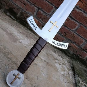 May include: A medieval sword with a silver blade and a brown leather-wrapped handle. The crossguard features a gold cross and the word "CHRISTUS". The pommel also has a gold cross. The word "VINCIT" is inscribed on the blade.