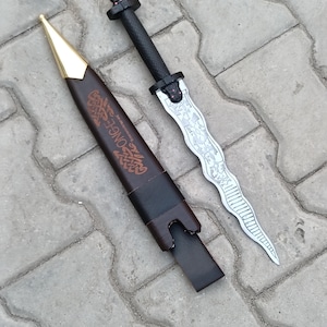May include: A decorative dagger with a silver blade and black handle, next to a brown leather sheath with gold accents. The blade has an ornate design. The sheath has the word "ONGE" printed on it.