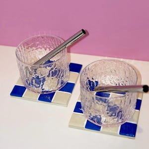Puede incluir: Dos vasos de vidrio transparente texturizado con pajitas de acero inoxidable descansan sobre posavasos cuadrados con un patrón de cuadros azules y blancos. El fondo es rosa liso.