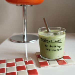 May include: A glass of green beverage with a straw, resting on a red and white checkered coaster. The glass has handwritten text that reads "Today's List: Be fucking incredible." An orange lamp is in the background.