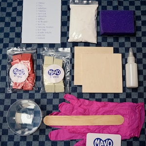 Könnte beinhalten: Ein Bastelset mit verschiedenen Materialien, darunter ein Beutel mit Pulver, ein lila Schwamm, Holzbretter, eine Flasche und rosa Handschuhe. Das Set enthält auch kleine Beutel mit dem Logo "MAVO STUDIO" und eine Liste mit der Aufschrift "HAPPY".