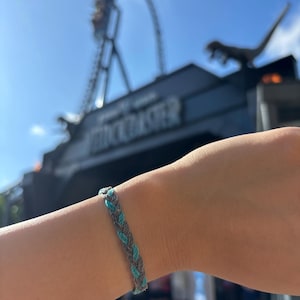 May include: Braided bracelet with alternating strands of gray and teal blue. The bracelet is worn on a wrist, with a blurred background of a roller coaster and a sign that says VELOCICOASTER.