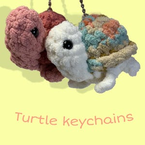 May include: Three crocheted turtle keychains in shades of pink, white, and multicolored yarn. Each turtle has a black eye and is attached to a silver keychain. The text "Turtle keychains" is written in pink.