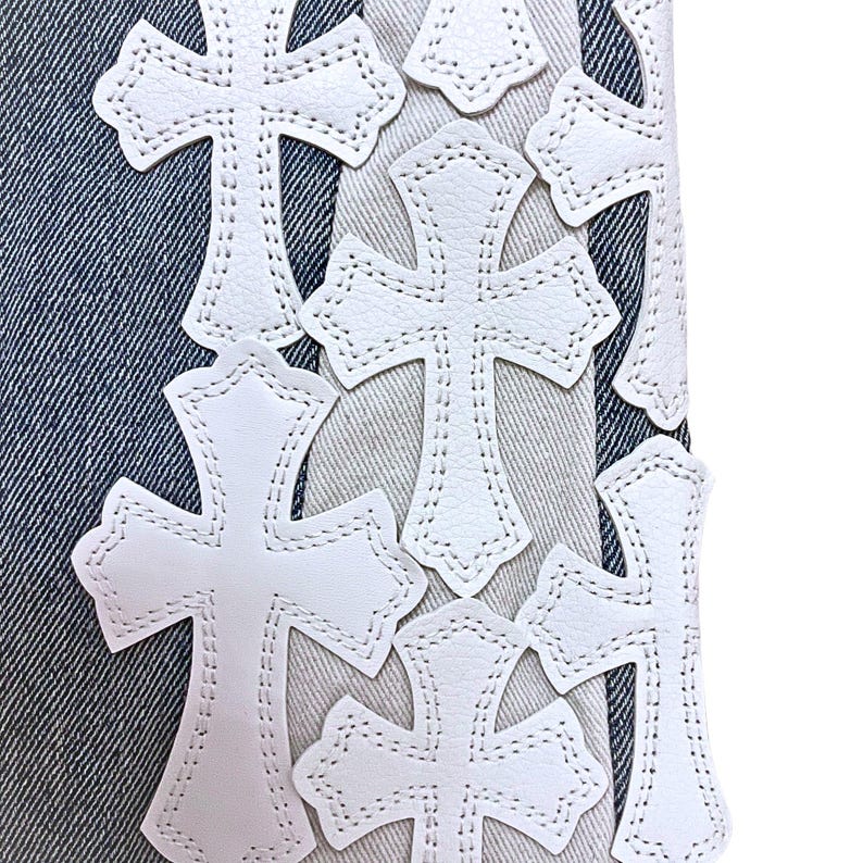 May include: Close-up of denim fabric with multiple white cross-shaped patches. The crosses are made of a smooth material and are stitched onto the fabric. The denim has a light blue wash.