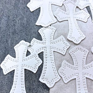 May include: White leather crosses with stitched edges are scattered across a denim background. The crosses have a detailed design and a textured surface. The image showcases a collection of decorative craft supplies.