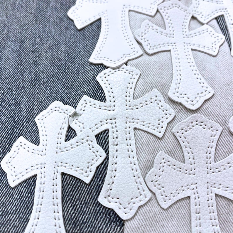 May include: White leather crosses with stitched edges are scattered across a denim background. The crosses have a detailed design and a textured surface. The image showcases a collection of decorative craft supplies.