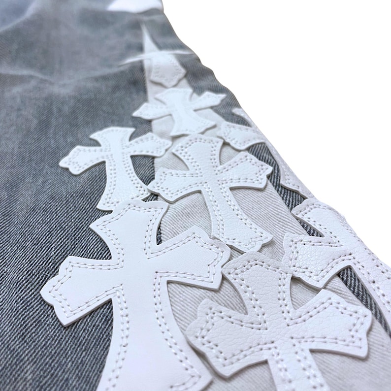 May include: Close-up of denim fabric with white leather cross appliques. The crosses are stitched onto the fabric, creating a textured and decorative design. The denim has a faded, worn appearance.