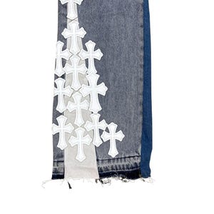 May include: Close-up of denim jeans with white cross appliques. The jeans have a distressed look with frayed edges and a mix of light and dark blue denim. The crosses are evenly spaced along the side.