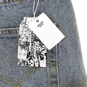 May include: Close-up of blue denim jeans with yellow stitching. A white tag with "GALLERY DEPT." is attached. Another tag features a collage of words including "ARTH CRISIS" and "COMPUTER CRIME".