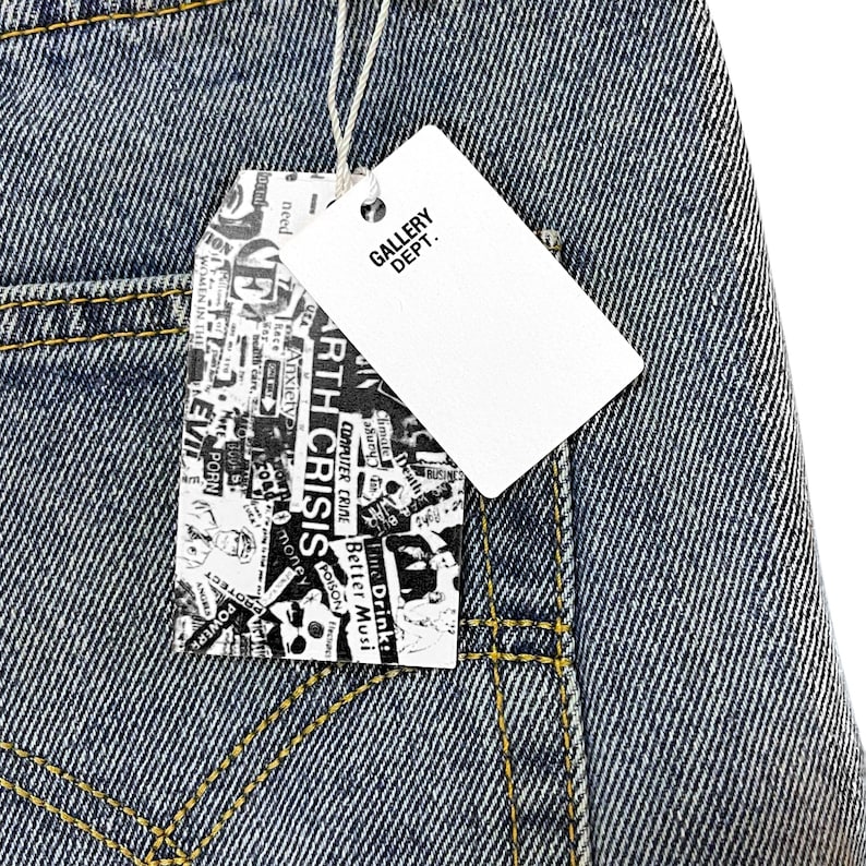 May include: Close-up of blue denim jeans with yellow stitching. A white tag with "GALLERY DEPT." is attached. Another tag features a collage of words including "ARTH CRISIS" and "COMPUTER CRIME".