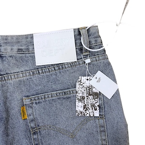 May include: Faded blue denim jeans with a white leather tag embossed with "GALLERY DEPT." and a yellow tag with "GALLERY DEPT." on the back pocket. A price tag with the text "EARTH CRISIS" is attached.