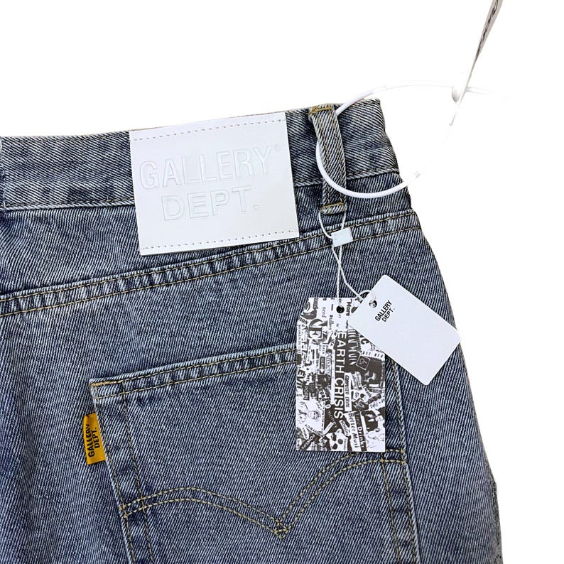 May include: Faded blue denim jeans with a white leather tag embossed with "GALLERY DEPT." and a yellow tag with "GALLERY DEPT." on the back pocket. A price tag with the text "EARTH CRISIS" is attached.