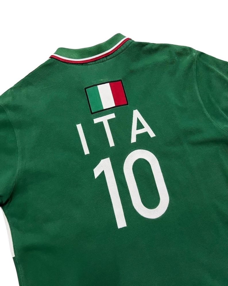 Pu&ograve; includere: Polo verde con la toppa della bandiera italiana, le lettere "ITA" e il numero "10" in bianco. Il colletto ha strisce rosse e bianche. La maglia &egrave; realizzata in materiale morbido.