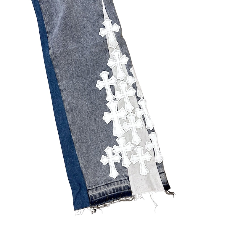 May include: Close-up of denim jeans with a unique design. The jeans feature a dark wash on the front and a contrasting blue panel on the side. White cross appliques adorn the right leg, adding a distinctive touch to the garment.