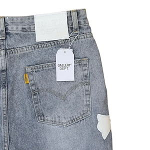 May include: Faded blue denim jeans with a back pocket and a white patch. The jeans have a white tag that says "GALLERY DEPT." and a yellow tag on the back pocket. The jeans are a light wash.