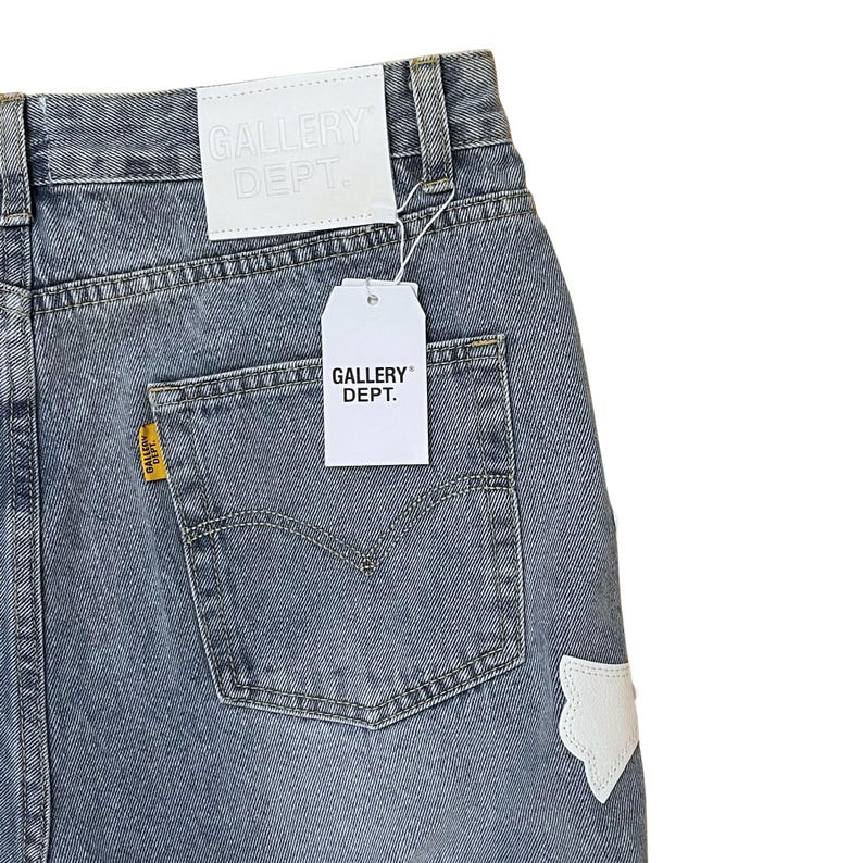 May include: Faded blue denim jeans with a back pocket and a white patch. The jeans have a white tag that says "GALLERY DEPT." and a yellow tag on the back pocket. The jeans are a light wash.