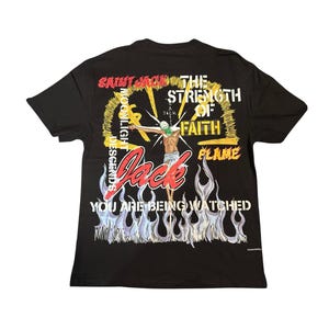 May include: Black t-shirt with a graphic design featuring a figure with outstretched arms, a green mask, and the word "Jack" in red. The design includes flames, text, and a yellow halo.