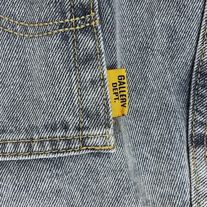 May include: Close-up of denim fabric with yellow stitching and a yellow tag. The tag reads "GALLERY DEPT." in black capital letters. The denim has a faded, worn appearance.