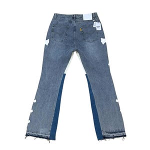May include: Faded blue denim jeans with white and dark blue accents. The jeans have a classic five-pocket design, a frayed hem, and white patches. A tag is attached to the back pocket.