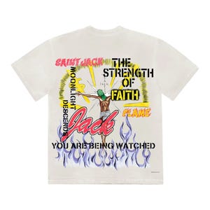 May include: Cream-colored t-shirt with a graphic design featuring the text "SAINT JACK" and "THE STRENGTH OF FAITH." The design includes an illustration of a figure with a green mask and the words "YOU ARE BEING WATCHED."