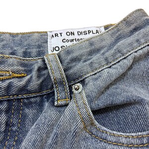 May include: Close-up of blue denim jeans with a button and pocket detail. The jeans have yellow stitching and a white tag that reads "ART ON DISPLAY" and "JOS". The denim has a faded, worn look.