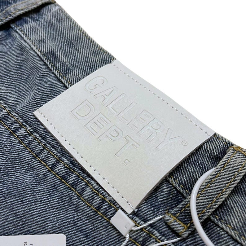 May include: Close-up of denim jeans with a white leather tag. The tag is embossed with the words "GALLERY DEPT." in capital letters. The jeans have a light blue wash and yellow stitching.