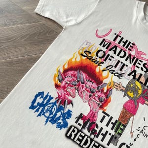 May include: White t-shirt with a graphic design featuring the text "THE MADNESS OF IT ALL" and "CHAOS". The design includes a three-headed pink monster with flames and other illustrations. The text "Saint Jack" is also visible.