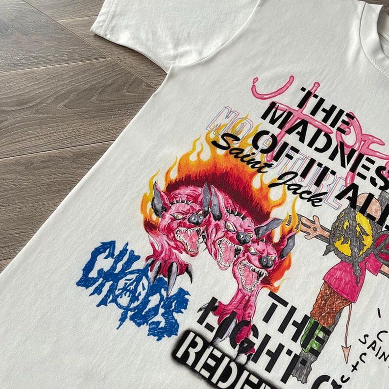 May include: White t-shirt with a graphic design featuring the text "THE MADNESS OF IT ALL" and "CHAOS". The design includes a three-headed pink monster with flames and other illustrations. The text "Saint Jack" is also visible.