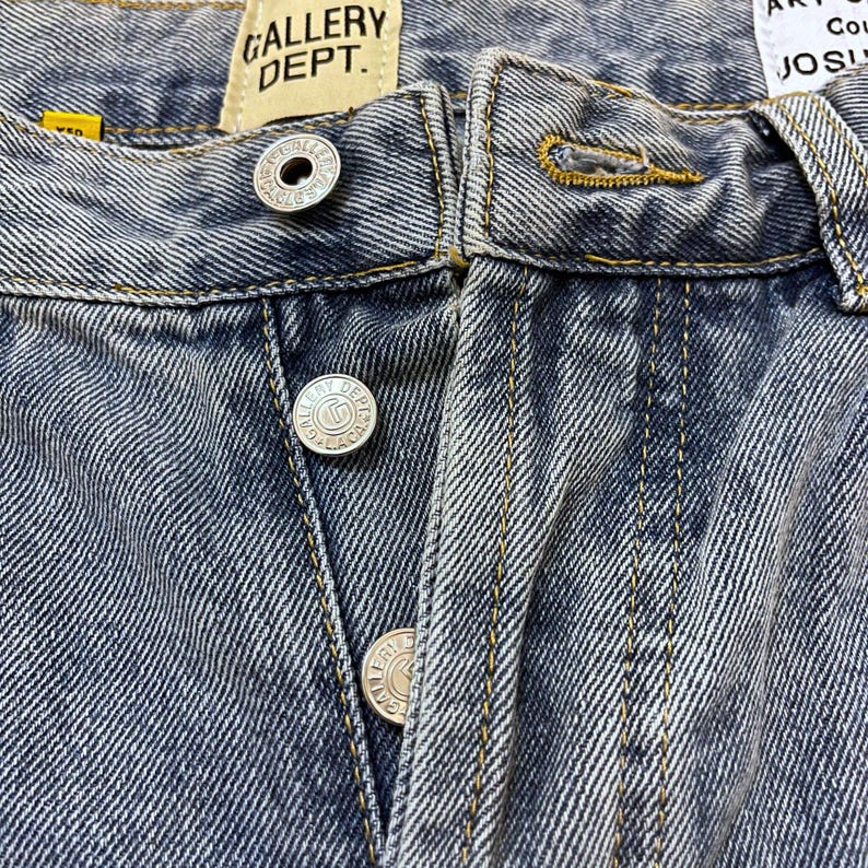 May include: Close-up of blue denim jeans with visible stitching and silver button closures. The jeans feature the "GALLERY DEPT." label. The denim has a faded, worn appearance.