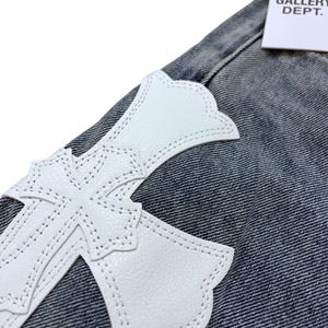 May include: Close-up of denim fabric with a large white cross-shaped leather patch. The cross is stitched onto the denim. A white tag with the text "GALLERY DEPT." is visible in the upper right corner.