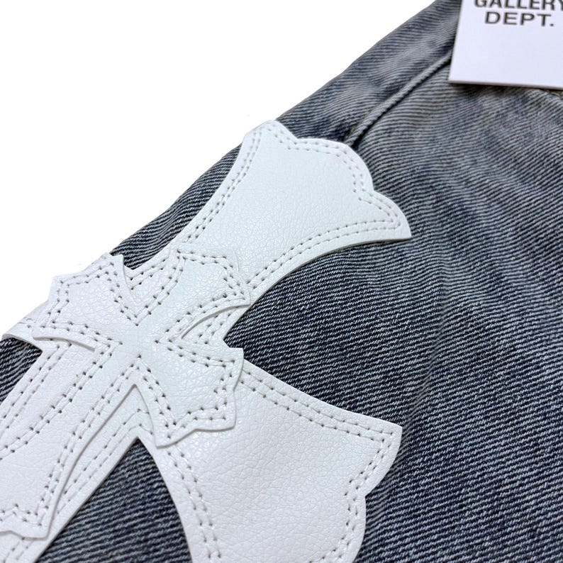May include: Close-up of denim fabric with a large white cross-shaped leather patch. The cross is stitched onto the denim. A white tag with the text "GALLERY DEPT." is visible in the upper right corner.