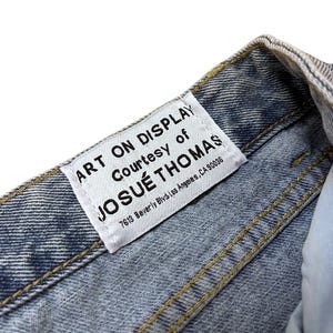 May include: Close-up of a light blue denim garment with yellow stitching. A white label is sewn into the garment, with the text "ART ON DISPLAY Courtesy of JOSU&Eacute; THOMAS 7613 Beverly Blvd Los Angeles, CA 90036".