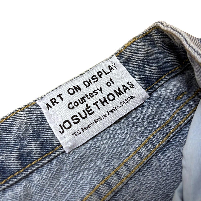May include: Close-up of a light blue denim garment with yellow stitching. A white label is sewn into the garment, with the text "ART ON DISPLAY Courtesy of JOSU&Eacute; THOMAS 7613 Beverly Blvd Los Angeles, CA 90036".