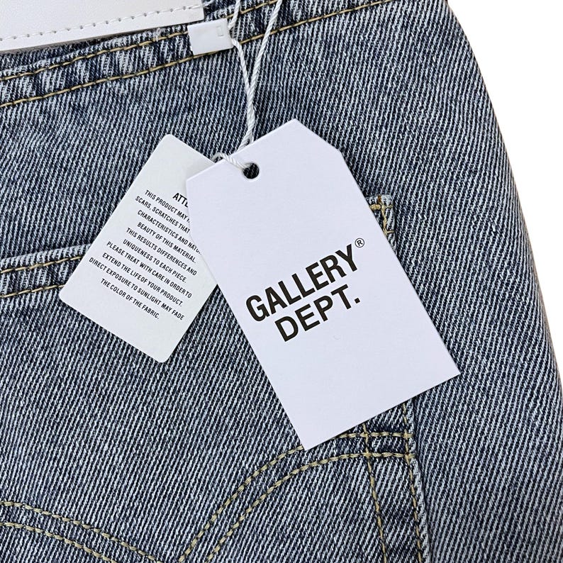 May include: Close-up of denim fabric with a white tag that reads "GALLERY DEPT." in black block letters. A smaller tag with text is also visible. The denim has visible stitching and a faded appearance.