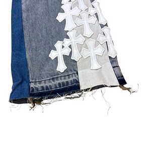 May include: Close-up of denim jeans with a patchwork design. The jeans feature a mix of blue, gray, and white denim panels. White cross-shaped patches are sewn onto the gray panel. The hem is frayed.