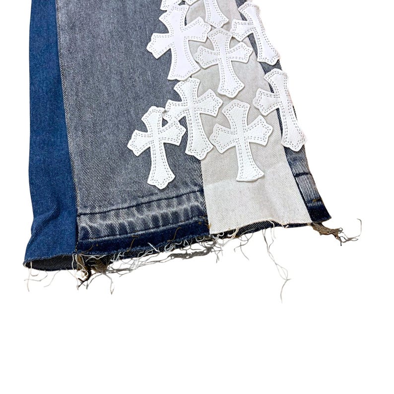 May include: Close-up of denim jeans with a patchwork design. The jeans feature a mix of blue, gray, and white denim panels. White cross-shaped patches are sewn onto the gray panel. The hem is frayed.