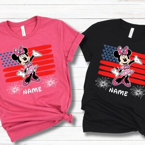 May include: Two t-shirts, one pink and one black, featuring a Minnie Mouse graphic. The design includes a stylized American flag with a Minnie Mouse character, fireworks, and the word "NAME" at the bottom. The shirts are short-sleeved.