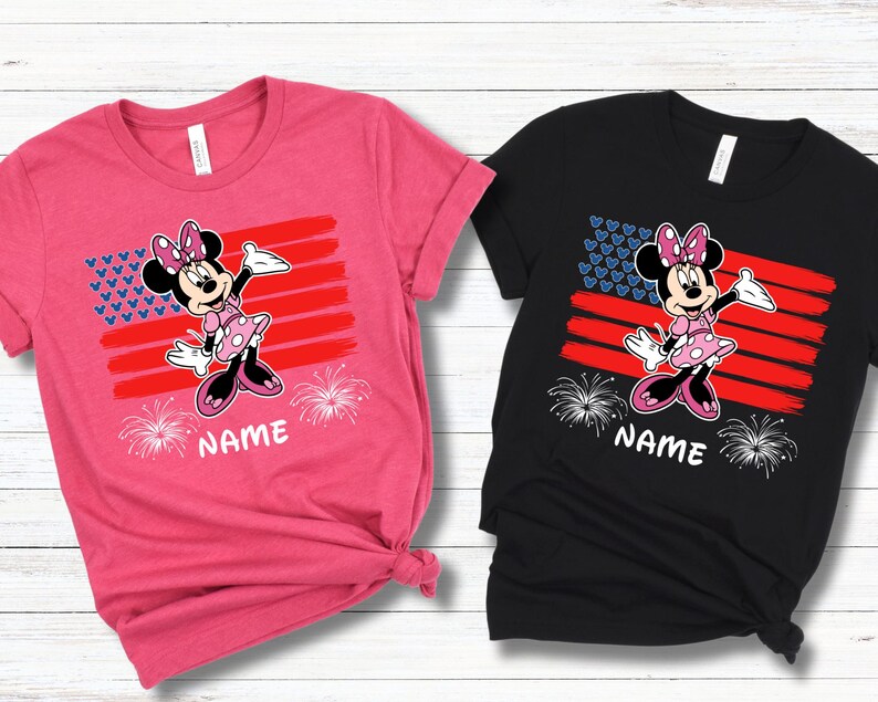 May include: Two t-shirts, one pink and one black, featuring a Minnie Mouse graphic. The design includes a stylized American flag with a Minnie Mouse character, fireworks, and the word "NAME" at the bottom. The shirts are short-sleeved.
