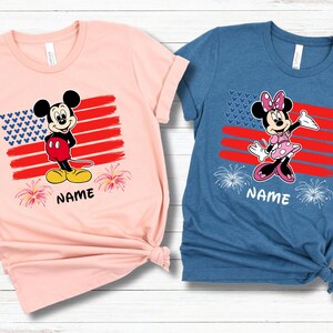 May include: Two short-sleeved t-shirts, one pink and one blue, featuring cartoon characters. Each shirt has a design of an American flag with a character and the word "NAME" below. The pink shirt has Mickey Mouse, and the blue shirt has Minnie Mouse.