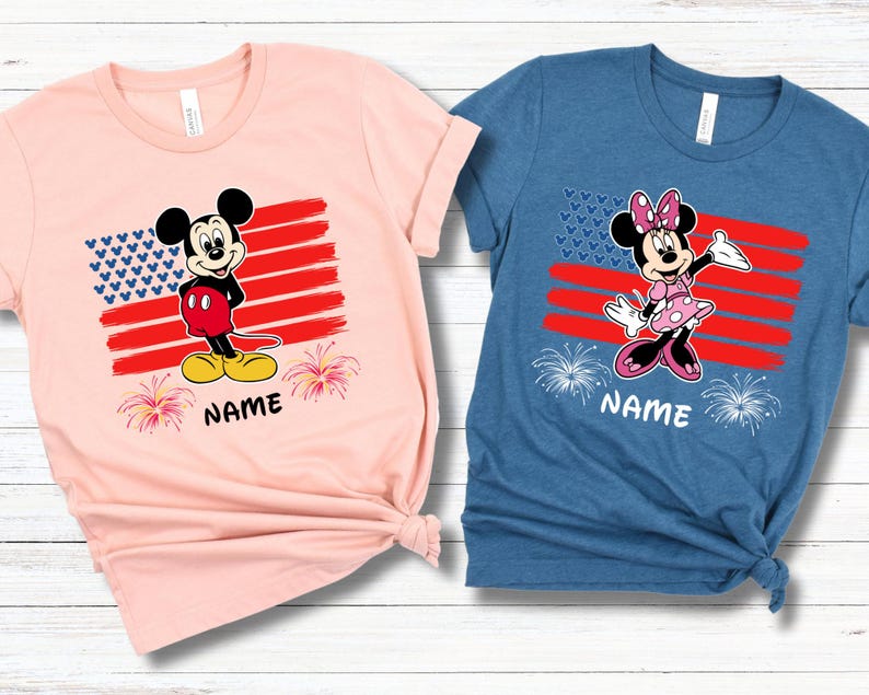 May include: Two short-sleeved t-shirts, one pink and one blue, featuring cartoon characters. Each shirt has a design of an American flag with a character and the word "NAME" below. The pink shirt has Mickey Mouse, and the blue shirt has Minnie Mouse.