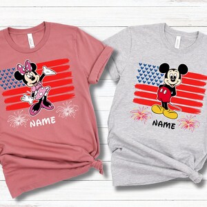 May include: Two short-sleeved t-shirts, one dusty rose and one heather gray. Each shirt features a cartoon character design with an American flag motif, fireworks, and the word "NAME" printed below the design.