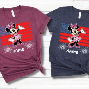May include: Two short-sleeved t-shirts, one burgundy and one navy blue, each featuring a Minnie Mouse graphic. The design includes a red and blue flag, fireworks, and the word "NAME" in white. The shirts are made of a soft material.