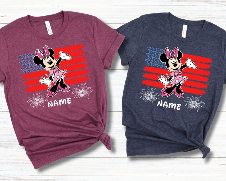 May include: Two short-sleeved t-shirts, one burgundy and one navy blue, each featuring a Minnie Mouse graphic. The design includes a red and blue flag, fireworks, and the word "NAME" in white. The shirts are made of a soft material.