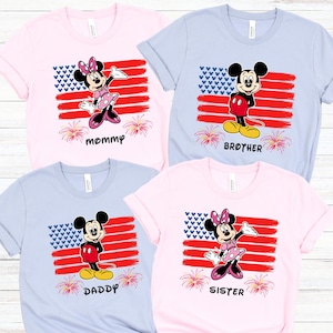 May include: Four short-sleeved t-shirts in pink and light blue, each featuring a cartoon character design. The designs include Mickey and Minnie Mouse in front of a stylized American flag, with the words "mommy", "brother", "daddy", and "sister" below.